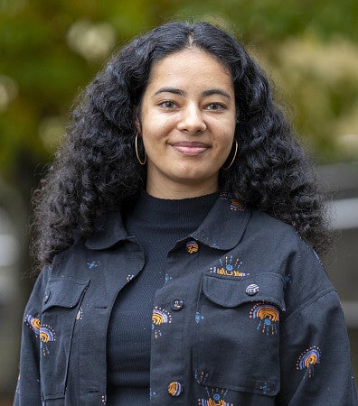 Portrait of Nayantara Arora looking at camera with long curly hair, hoop earrings and wearing a black denim jacket with embroidered designs